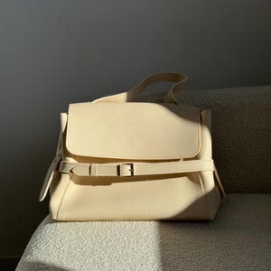 May include: A cream-colored handbag with a textured surface, featuring a fold-over flap and a horizontal strap across the front secured by a metal buckle. The bag includes a single top handle and side strap details, presented in a structured design.