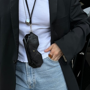 May include: A black leather glasses case with a gold clasp hangs from a silver chain necklace. The case is worn by a person wearing a white tank top and a black blazer. The person is wearing blue jeans and a gold ring.