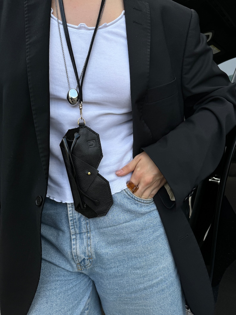 May include: A black leather glasses case with a gold clasp hangs from a silver chain necklace. The case is worn by a person wearing a white tank top and a black blazer. The person is wearing blue jeans and a gold ring.