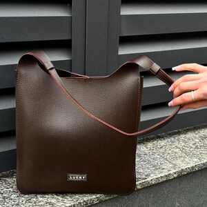 May include: A brown leather tote bag with a rectangular shape and a shoulder strap. The bag has a textured surface and a small metal tag that reads "LUCKY". The bag is sitting on a stone surface.
