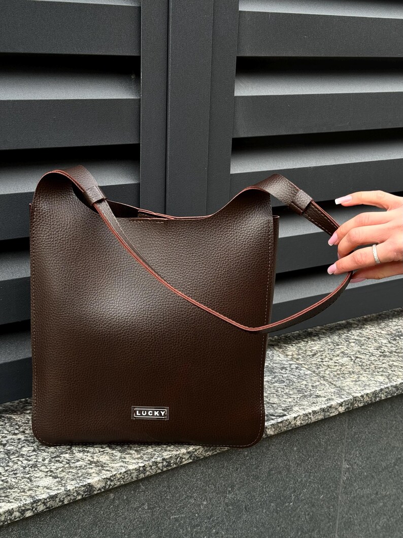 May include: A brown leather tote bag with a rectangular shape and a shoulder strap. The bag has a textured surface and a small metal tag that reads "LUCKY". The bag is sitting on a stone surface.