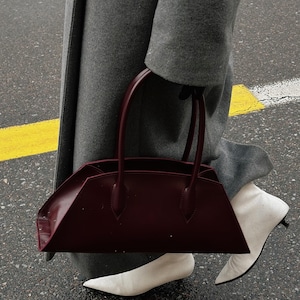 May include: A burgundy leather handbag with a structured, angular design. The bag has two top handles and is held by a person wearing a gray coat and white pointed-toe boots. The background shows a wet street with a yellow stripe.