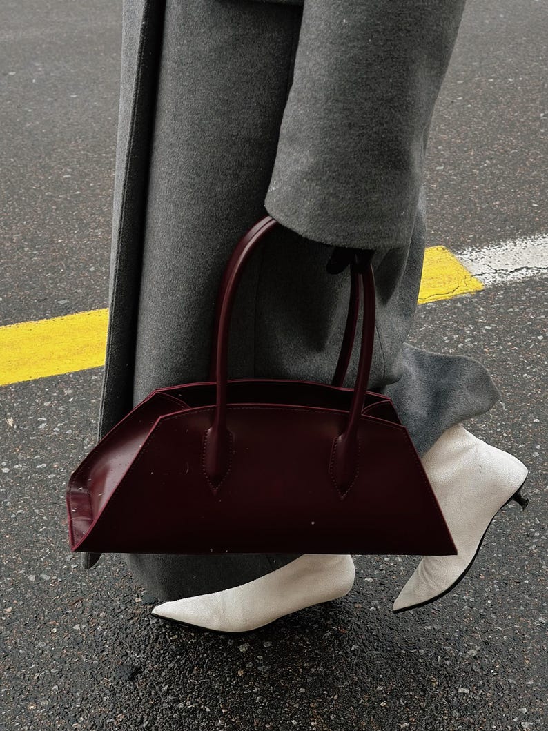 May include: A burgundy leather handbag with a structured, angular design. The bag has two top handles and is held by a person wearing a gray coat and white pointed-toe boots. The background shows a wet street with a yellow stripe.