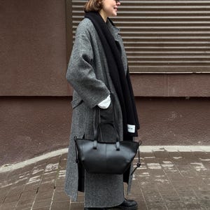 May include: A black leather tote bag with a top handle and a shoulder strap. The bag is held by a person wearing a gray wool coat, a black scarf, and a black hat. The background is a brown wall and a brick sidewalk.