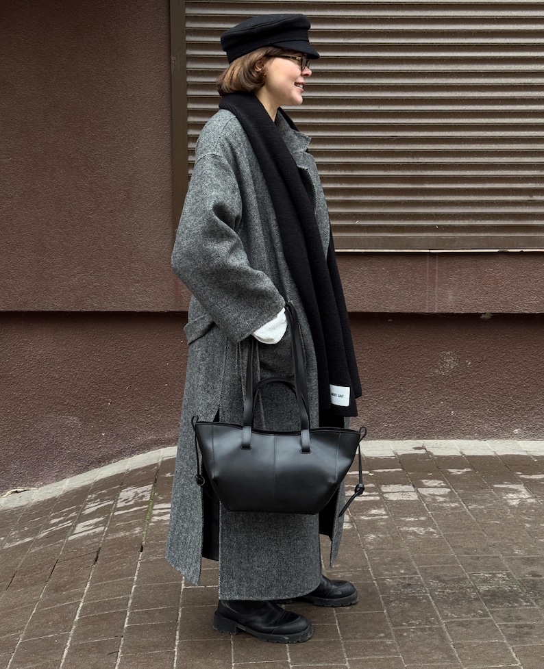 May include: A black leather tote bag with a top handle and a shoulder strap. The bag is held by a person wearing a gray wool coat, a black scarf, and a black hat. The background is a brown wall and a brick sidewalk.