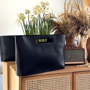 Könnte beinhalten: Eine elegante schwarze Ledertasche mit rechteckiger Silhouette und einem in die Oberkante integrierten Griffausschnitt. Die Tasche hat eine glatte, matte Oberfläche und ein strukturiertes Design, dargestellt als ein Paar identischer Stücke.