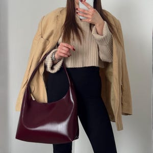 Op de afbeelding: Een gladde, diepbordeauxrode hobo-schoudertas met een enkele riem en goudkleurige hardware. De tas heeft een gebogen silhouet en een gepolijste afwerking, gedragen bij een beige geribbelde trui, een beige blazer en een zwarte broek.