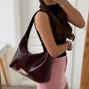 Peut inclure: Un sac à bandoulière de style hobo bordeaux foncé avec une finition lisse et semi-brillante. Le sac présente une seule bandoulière intégrée, une ouverture supérieure incurvée et un design minimaliste aux lignes épurées sans poches.