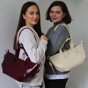 May include: Two women holding tote bags. One bag is a glossy burgundy color, and the other is a cream color. Both bags have short handles and a rectangular shape. The women are standing in front of a plain white wall.