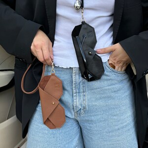 May include: Two leather eyeglass cases, one brown and one black, are hanging from a silver chain around a person's neck. The cases are folded and have a geometric shape. The person is wearing a white shirt and blue denim shorts.