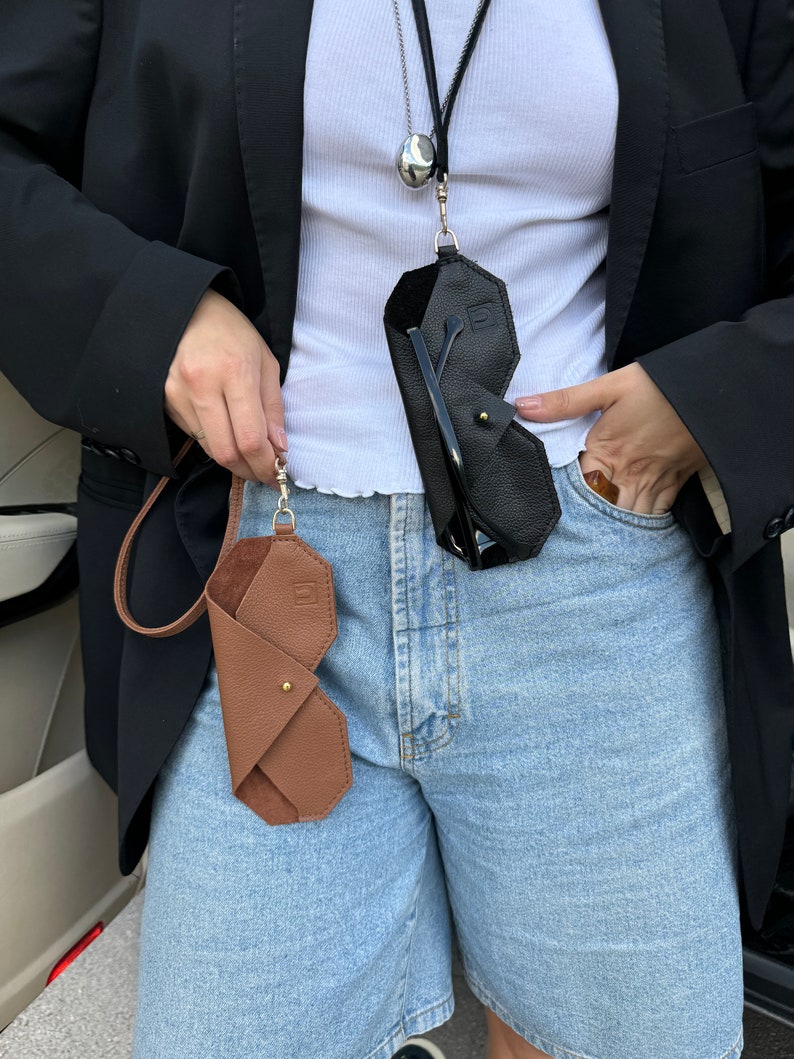 May include: Two leather eyeglass cases, one brown and one black, are hanging from a silver chain around a person's neck. The cases are folded and have a geometric shape. The person is wearing a white shirt and blue denim shorts.