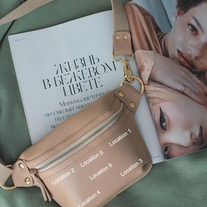 May include: A beige leather fanny pack with a gold zipper and buckle. The fanny pack has six numbered locations on the front.