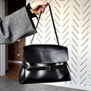 May include: A black handbag with a smooth finish, featuring a top flap closure and a horizontal strap across the front secured by a silver-toned metal buckle. The bag includes a single thin shoulder strap attached to the top sides of the main body.