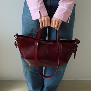 May include: A burgundy leather tote bag with a structured design. The bag features a top zipper closure, a long shoulder strap, and two short handles. The bag is held by a person wearing blue jeans and a pink shirt.