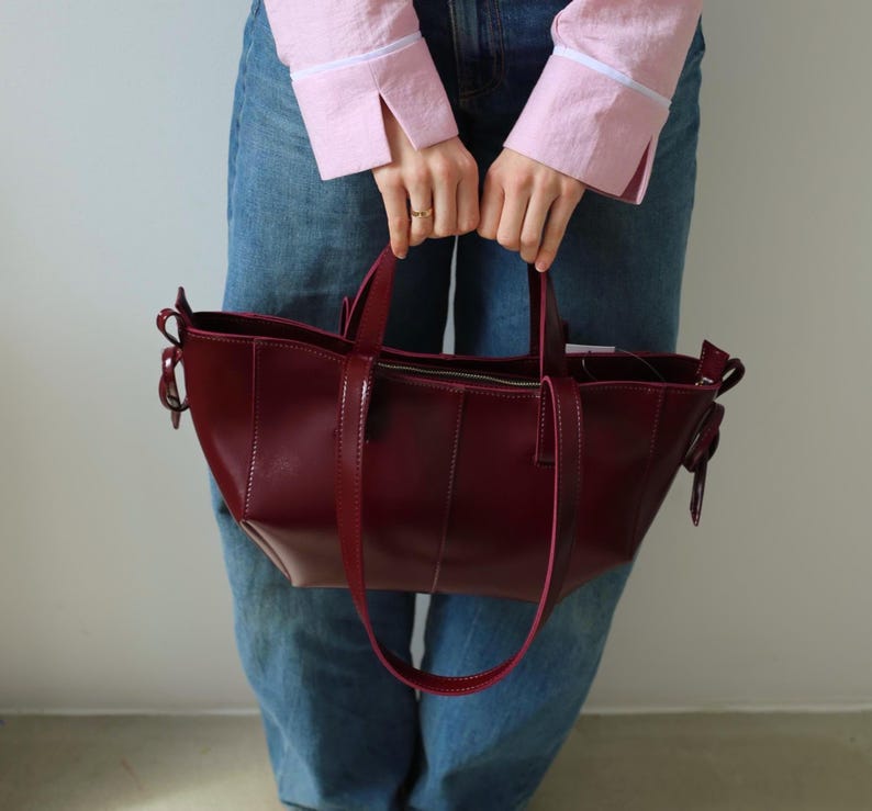 May include: A burgundy leather tote bag with a structured design. The bag features a top zipper closure, a long shoulder strap, and two short handles. The bag is held by a person wearing blue jeans and a pink shirt.