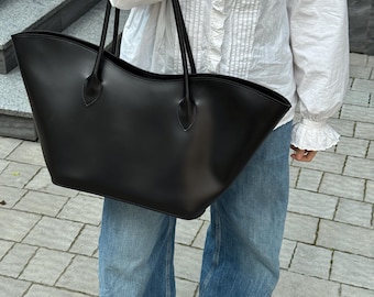May include: A large, smooth black tote bag with a wide, curved top opening and two thin shoulder straps. The bag features a structured base and a minimalist design, crafted from a solid black material with a subtle sheen, held by the straps.