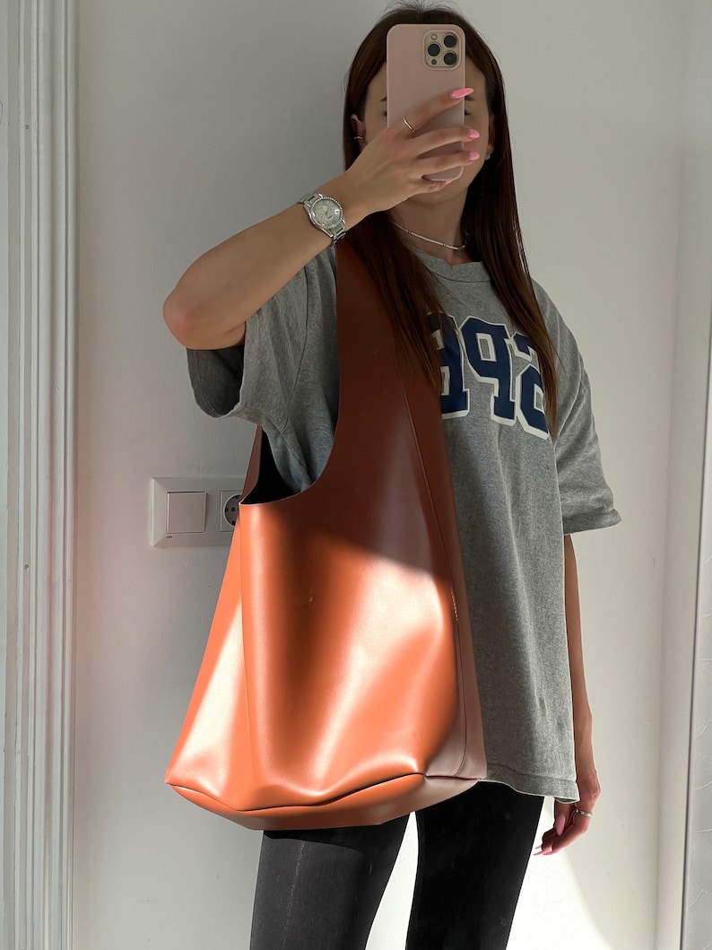 May include: A brown leather tote bag with a wide, open top. The bag is being held by a person wearing a grey t-shirt with the word "94" printed on it.