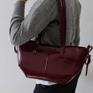 May include: A burgundy leather tote bag with a rectangular shape and a glossy finish. The bag has two handles and a zipper closure. The bag is being held by a person wearing a gray sweater.