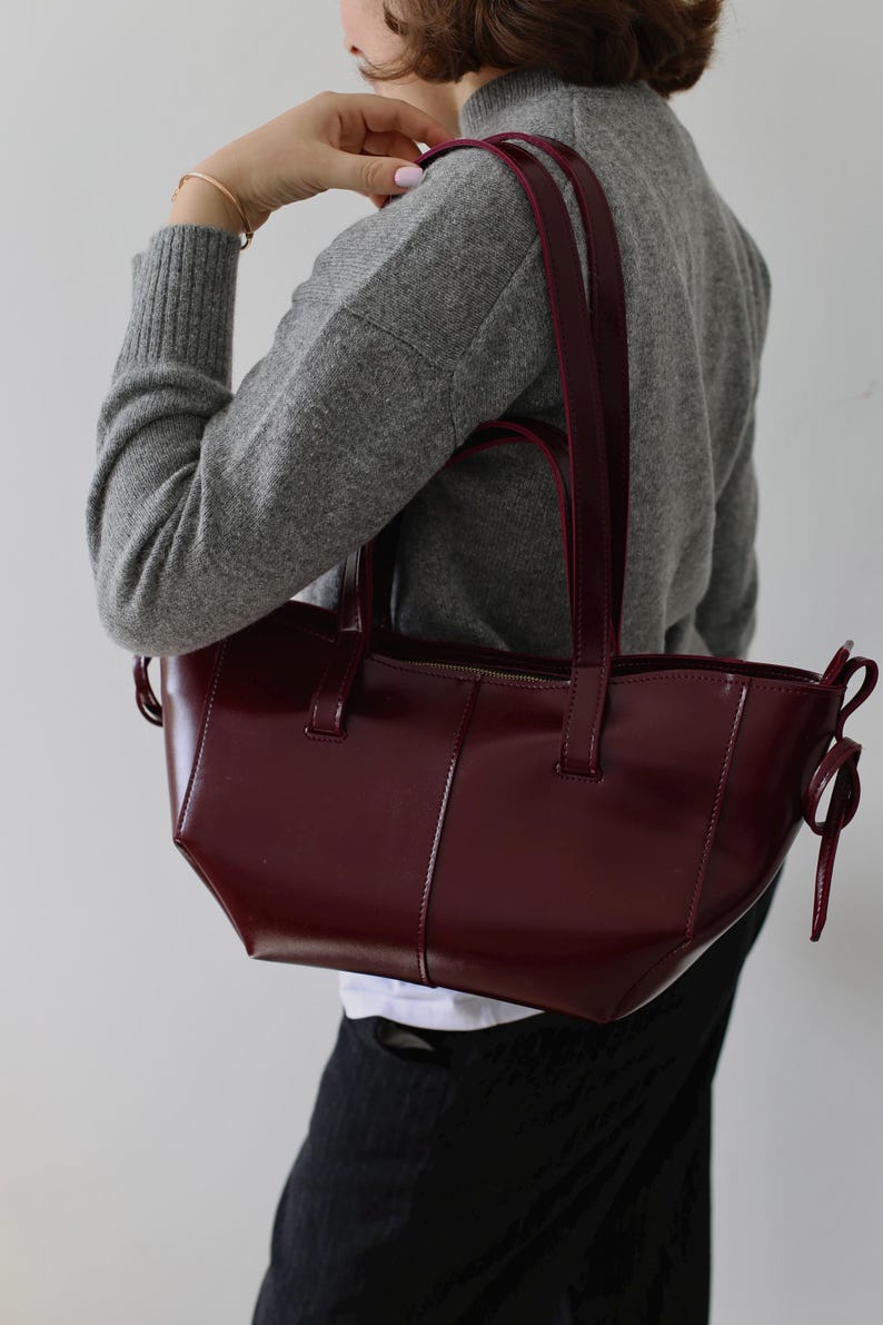 May include: A burgundy leather tote bag with a rectangular shape and a glossy finish. The bag has two handles and a zipper closure. The bag is being held by a person wearing a gray sweater.