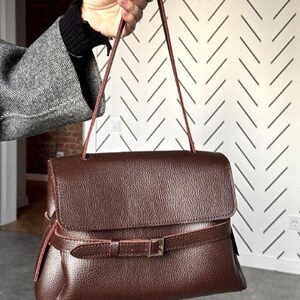 May include: A dark brown handbag with a textured, pebbled surface, featuring a fold-over flap and a thin, adjustable strap with a silver-toned metal buckle closure that wraps horizontally around the lower section of the bag body.