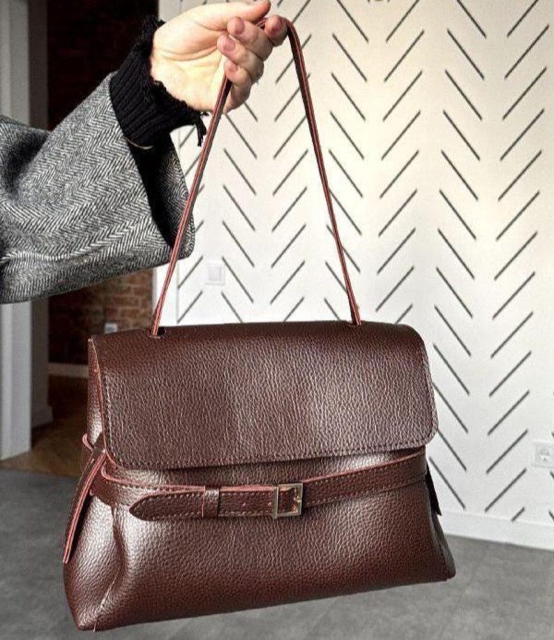 May include: A dark brown handbag with a textured, pebbled surface, featuring a fold-over flap and a thin, adjustable strap with a silver-toned metal buckle closure that wraps horizontally around the lower section of the bag body.