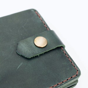 May include: Close-up of a dark green leather wallet with a brown stitch detail and a gold button closure. The wallet is made of genuine leather and is hand-made.