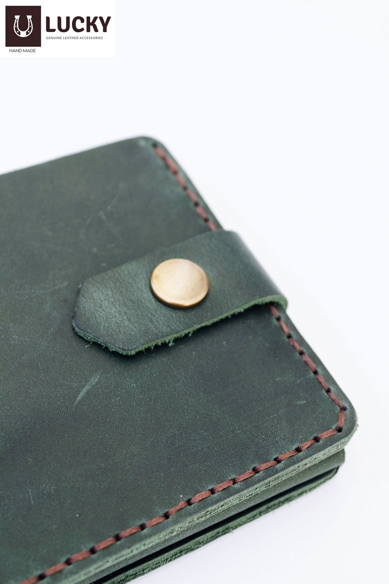 May include: Close-up of a dark green leather wallet with a brown stitch detail and a gold button closure. The wallet is made of genuine leather and is hand-made.