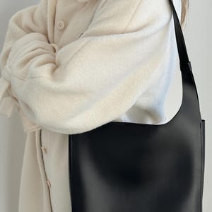 May include: A black shoulder bag with a rectangular shape and a long strap. The bag has a minimalist design with the word "LUCKY" printed on the front. The bag is held by a person wearing a cream-colored coat.