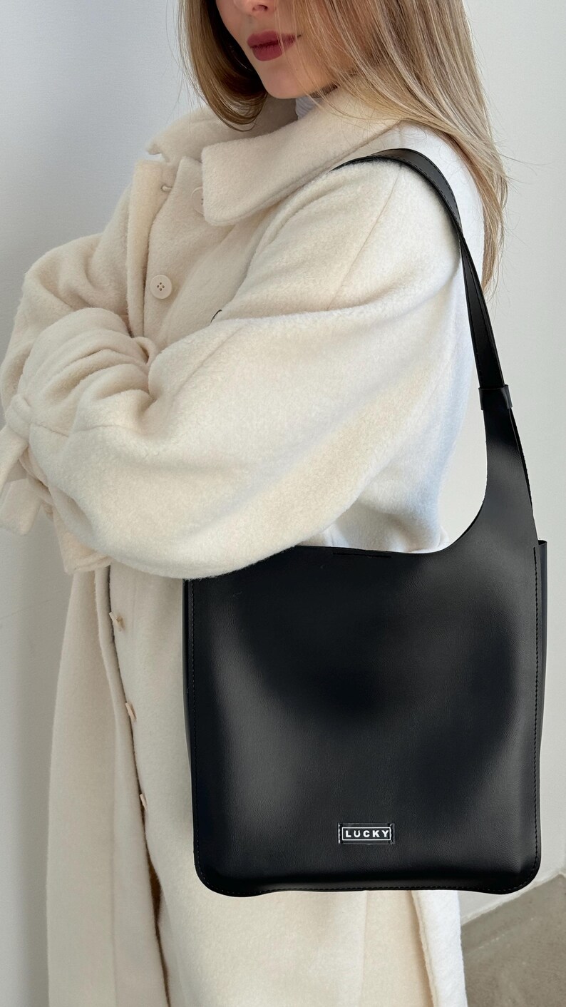 May include: A black shoulder bag with a rectangular shape and a long strap. The bag has a minimalist design with the word "LUCKY" printed on the front. The bag is held by a person wearing a cream-colored coat.