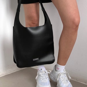May include: A black leather shoulder bag with a rectangular shape and a long strap. The bag has a small, rectangular silver label that reads "LUCKY". The person is wearing white sneakers and white socks.