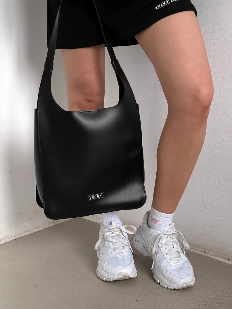 May include: A black leather shoulder bag with a rectangular shape and a long strap. The bag has a small, rectangular silver label that reads "LUCKY". The person is wearing white sneakers and white socks.