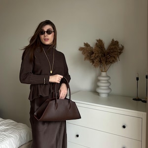 May include: A woman wearing a brown outfit, including a turtleneck sweater, skirt, and sunglasses, holds a dark brown handbag. The outfit is complemented by a gold necklace and bracelet. A white dresser and decorative vase are in the background.