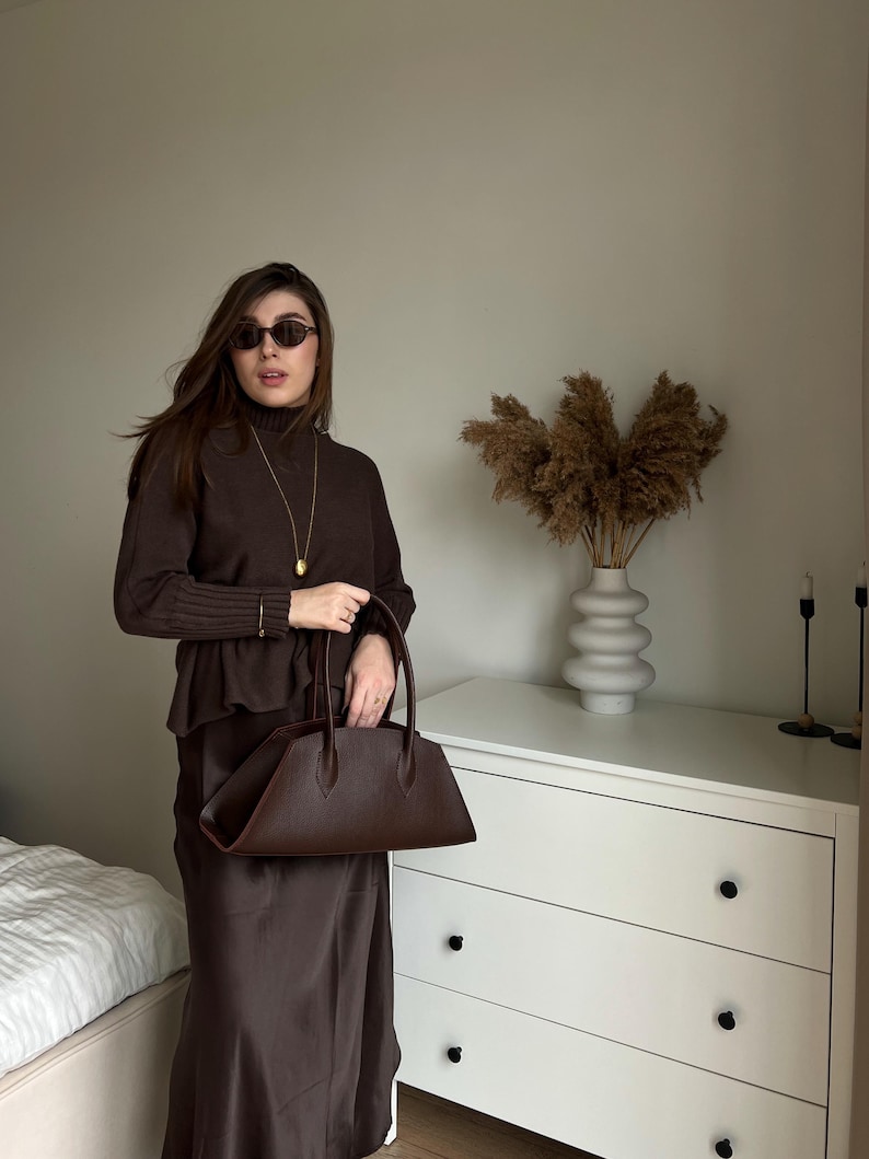 May include: A woman wearing a brown outfit, including a turtleneck sweater, skirt, and sunglasses, holds a dark brown handbag. The outfit is complemented by a gold necklace and bracelet. A white dresser and decorative vase are in the background.