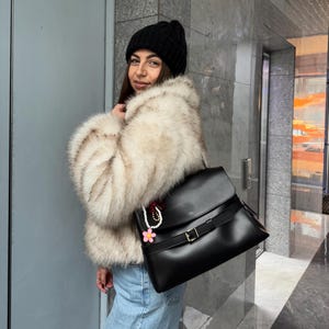 May include: A black leather satchel with a fold-over flap, a horizontal strap secured by a gold-tone buckle, and a decorative charm featuring a pink flower and white beads attached to the side. The bag is worn over the shoulder with a strap.