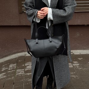 May include: A black leather tote bag with a structured design. The bag has two top handles and a shoulder strap. The person is wearing a gray coat, black scarf, and black boots.