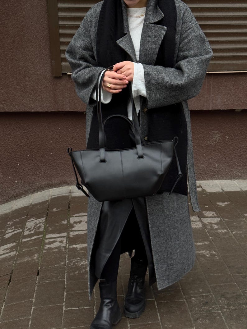 May include: A black leather tote bag with a structured design. The bag has two top handles and a shoulder strap. The person is wearing a gray coat, black scarf, and black boots.