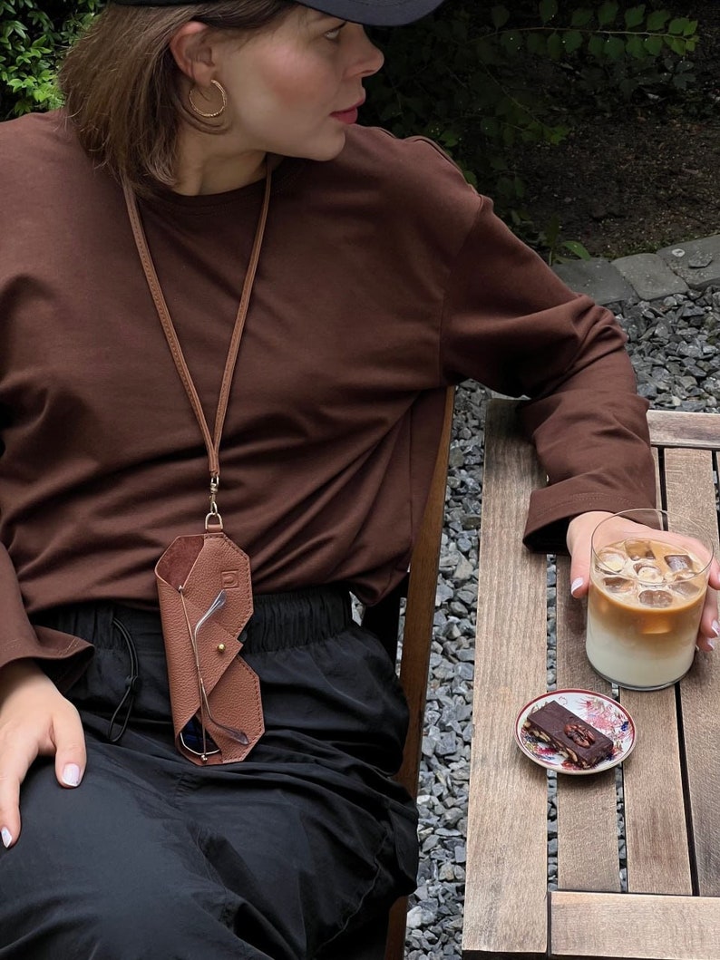 May include: A brown leather glasses case with a gold clasp hangs from a brown leather strap around a person's neck. The case is folded and has a flap that covers the glasses. The person is wearing a brown long-sleeved shirt and black pants.