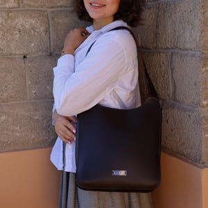 May include: A dark brown leather shoulder bag with a rectangular shape. The bag has a long strap and a small silver-colored metal tag on the front. The bag is being held by a person wearing a white shirt and a plaid skirt.
