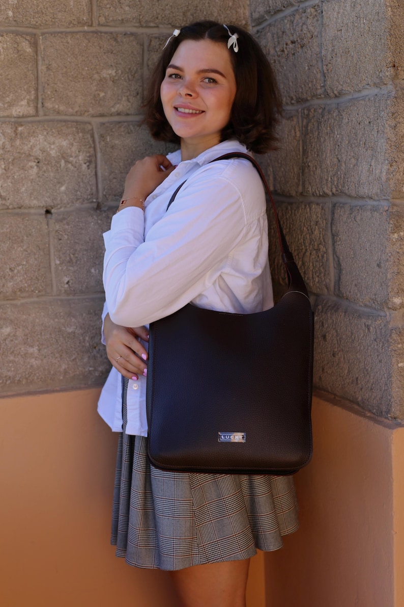 May include: A dark brown leather shoulder bag with a rectangular shape. The bag has a long strap and a small silver-colored metal tag on the front. The bag is being held by a person wearing a white shirt and a plaid skirt.