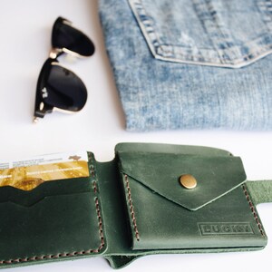 May include: A green leather wallet with a flap pocket and a brass button. The wallet has the word "LUCKY" embossed on the front. The wallet is open and shows a card slot and a billfold. The wallet is on a white surface with a pair of sunglasses and a piece of blue denim.