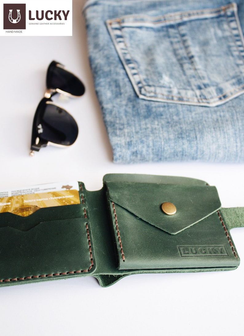 May include: A green leather wallet with a flap pocket and a brass button. The wallet has the word "LUCKY" embossed on the front. The wallet is open and shows a card slot and a billfold. The wallet is on a white surface with a pair of sunglasses and a piece of blue denim.