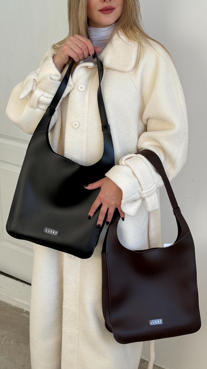 May include: Two shoulder bags, one black and one brown, are displayed. The black bag has a rectangular silver tag that reads "LUCKY". The brown bag has a similar tag. Both bags have a simple, minimalist design.