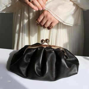 Peut inclure: Une pochette en cuir noir au design plissé et froncé, dotée d'une armature en métal doré. Le sac présente une fermeture à fermoir à cliquet avec deux sphères dorées polies et arrondies sur le dessus, assurant une ouverture décorative.