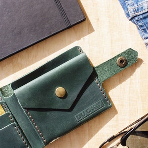 May include: A green leather wallet with a flap closure and a snap button. The wallet has a small pocket on the outside and the word "LUCKY" embossed on the front. The wallet is lying on a wooden surface with a notebook, sunglasses, and a pair of jeans.