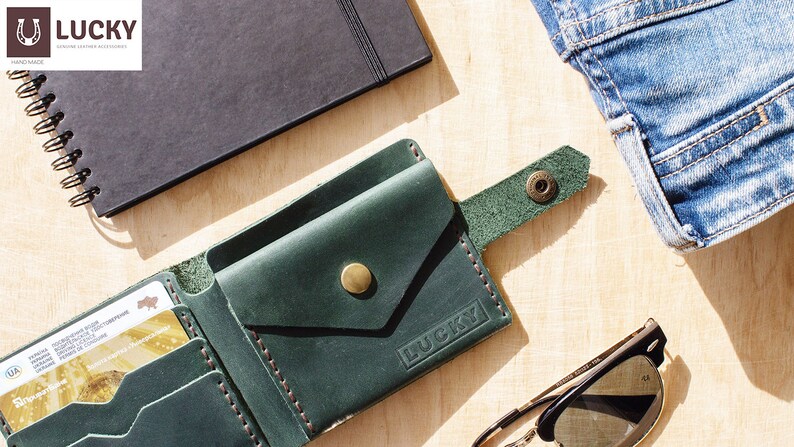 May include: A green leather wallet with a flap closure and a snap button. The wallet has a small pocket on the outside and the word "LUCKY" embossed on the front. The wallet is lying on a wooden surface with a notebook, sunglasses, and a pair of jeans.
