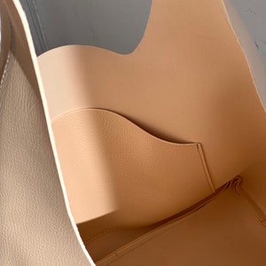 May include: A beige leather tote bag with a single interior pocket. The bag is open and the inside is visible.