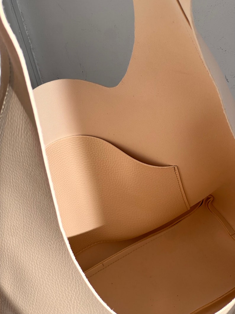 May include: A beige leather tote bag with a single interior pocket. The bag is open and the inside is visible.