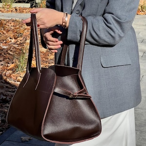 May include: A dark brown leather tote bag with a structured design. The bag features a top handle, a buckle closure, and a contrasting pink trim. The bag is held by a person wearing a gray blazer and a white skirt.