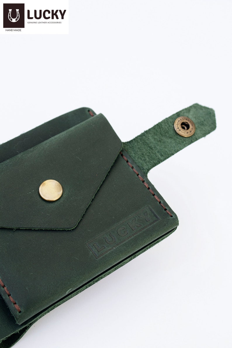May include: A dark green leather wallet with a flap pocket and a snap closure. The wallet has a brown stitched edge and the word "LUCKY" embossed on the front.