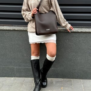 May include: A dark brown leather shoulder bag with a rectangular shape and a long strap. The bag is carried by a person wearing a beige sweater, white skirt, black boots, and white socks.